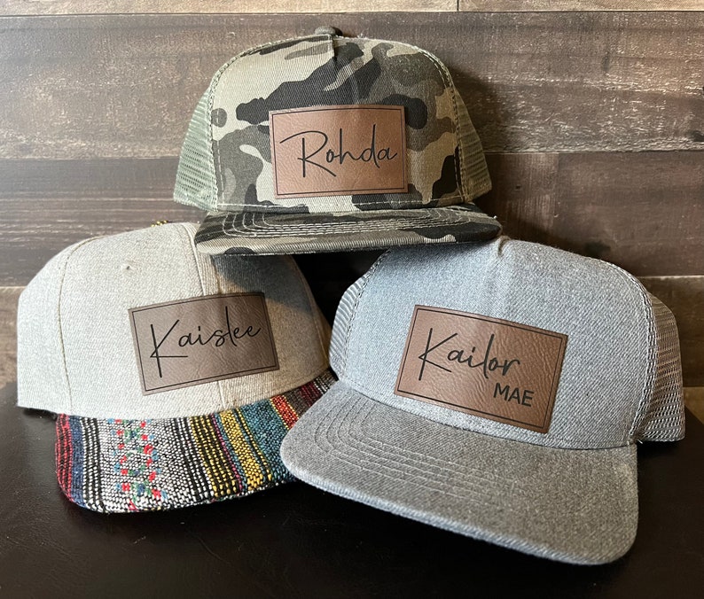 May include: Three gray and camo trucker hats with brown leather patches. The patches have the names "Rohda", "Kaislee", and "Kailor MAE" written in black cursive.
