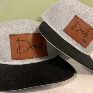 May include: Two gray baseball caps with black brims. The larger cap has a brown leather patch with the text "Dad's Mini" in black script. The smaller cap has a brown leather patch with the text "Dad" in black script.