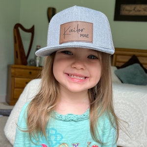 May include: A young girl wearing a gray and black trucker hat with a brown leather patch that says "Kailor MAE". She is wearing a teal shirt with a colorful "love" graphic.