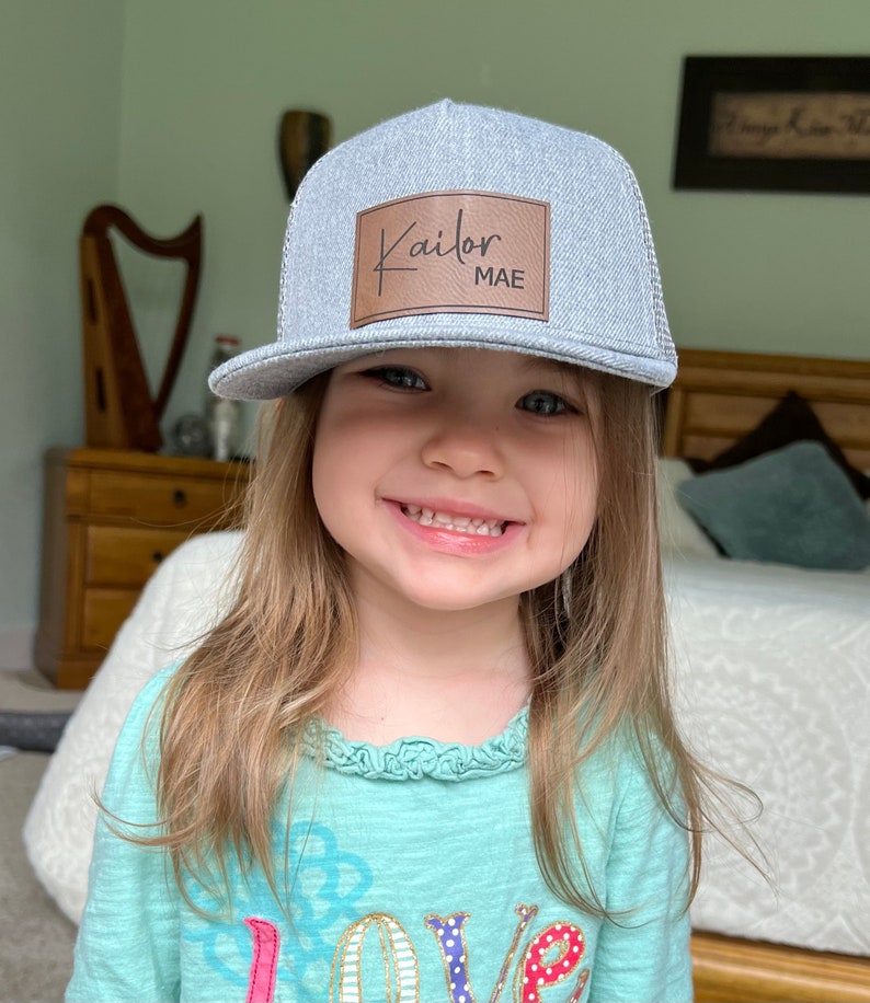 May include: A young girl wearing a gray and black trucker hat with a brown leather patch that says "Kailor MAE". She is wearing a teal shirt with a colorful "love" graphic.
