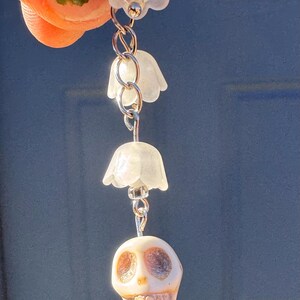 May include: A unique earring featuring a small, white skull pendant with silver-colored accents, suspended below three translucent, bell-shaped flower charms. The earring is attached to a silver-colored chain.