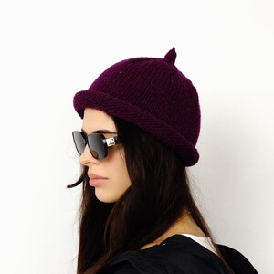 Puede incluir: Un gorro de punto morado oscuro con un borde pequeño y redondeado y un pequeño detalle puntiagudo en la parte superior. La persona lleva gafas de sol negras.