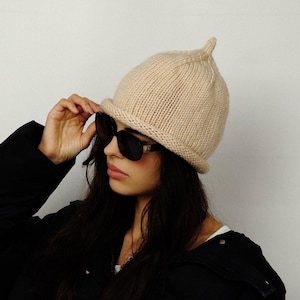 Peut inclure: Un bonnet beige en tricot avec un sommet pointu. Le chapeau est porté avec des lunettes de soleil noires et une veste noire. Le bonnet a un bord replié et un aspect texturé. Le style général est décontracté et moderne.