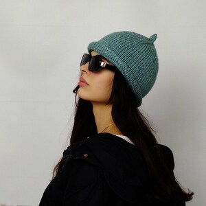 Puede incluir: Un gorro de punto color verde azulado con un borde pequeño y redondeado y un pequeño detalle puntiagudo en la parte superior. La persona lleva gafas de sol negras y una chaqueta negra.