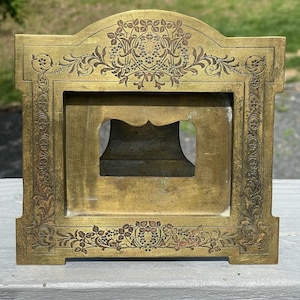 May include: A brass picture frame with an ornate design. The frame has a rectangular opening with a decorative border. The frame is made of solid brass and has a polished finish.