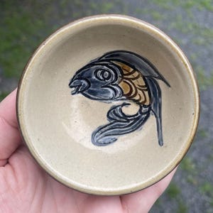 Puede incluir: Un pequeño cuenco de cerámica beige con un borde marrón. El cuenco tiene un diseño pintado a mano de un pez negro y dorado.