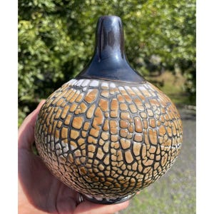May include: A ceramic vase with a distinctive textured pattern. It has a dark blue neck and a body with a mosaic-like design in shades of brown, white, and black. The vase is held in a hand.