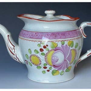 May include: White ceramic teapot with a lid, spout, and handle. The teapot is decorated with a pink band, floral designs in pink, yellow, and green, and red accents. The teapot has a curved spout and handle.