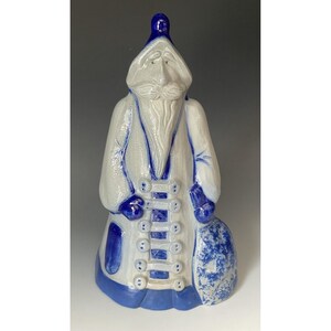 May include: A ceramic figurine of a bearded figure wearing a long coat and a pointed hat. The piece is glazed in white with deep blue accents on the hat, hem, buttons, and a sack held at the side. The figure has a long beard and moustache.
