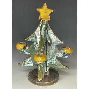 May include: A ceramic Christmas tree candle holder with a glossy green and white mottled glaze. The tree features a yellow star at the top and three circular yellow candle cups attached to the branches, all mounted on a dark brown circular base.