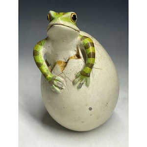 May include: A green and yellow frog figurine emerging from a cracked white egg. The frog has a textured surface and realistic details.