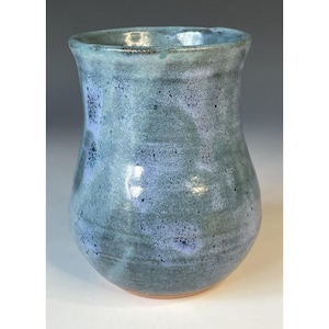 May include: A handmade ceramic vase with a speckled blue and lavender glaze. The vase has a rounded body and a slightly flared rim. The colors blend together, creating a unique and artistic piece. The base is a warm, earthy tone.