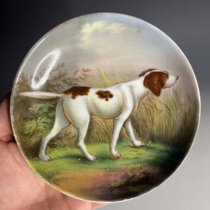 Puede incluir: Plato decorativo redondo con una pintura de un perro blanco y marrón en un campo. El perro es el punto focal, con una representación detallada de su pelaje y postura. El fondo incluye hierba, árboles y un cielo nublado, creando una escena pastoral.