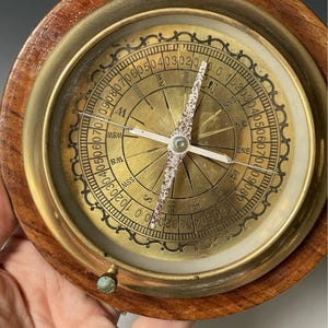 May include: A circular nautical compass featuring a polished brass housing set within a dark brown wooden frame. The face displays cardinal directions, degree markings, and a central needle with a textured, silver-toned finish and a small knob.