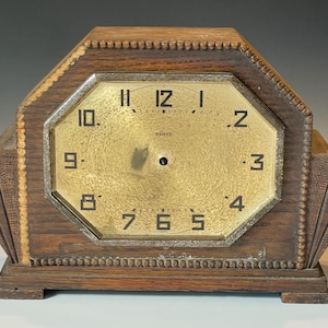 May include: Vintage wooden mantel clock with a gold-toned face and black numerals. The clock has an octagonal face and a rectangular wooden base. The clock face reads "8 DAYS". The clock's wooden frame has decorative detailing.