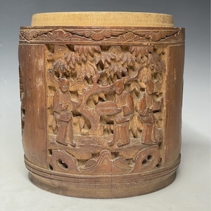 Peut inclure: Un récipient cylindrique en bois avec des sculptures complexes de personnages et d'arbres. Le récipient est en bois brun clair et a une finition lisse et polie.