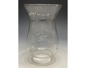 May include: Clear glass hurricane lamp shade with a flared top and etched leaf design. The shade has a rounded body and a circular base. The glass is transparent, allowing light to pass through. A decorative accent for home decor.