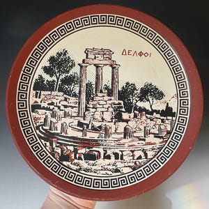May include: A round decorative plate featuring a black and cream illustration of the Tholos of Delphi ruins. The border has a black and white Greek key pattern on a terracotta red background. The Greek text "ΔΕΛΦΟΙ" is printed in red.