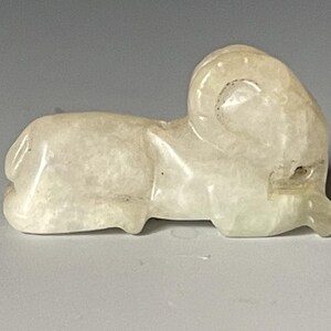 Könnte beinhalten: Kleine, cremefarbene Widderfigur aus durchscheinendem Material geschnitzt. Der Widder wird in Ruheposition dargestellt, mit seinen gekräuselten Hörnern und detaillierten Merkmalen. Die Skulptur ist glatt und poliert und zeigt die natürlichen Variationen im Stein.