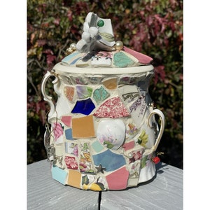 May include: A white ceramic jar with a mosaic design made from broken pieces of china and other materials. The jar has two handles and a lid with a white porcelain butterfly on top. The mosaic design features a variety of colors, including pink, blue, green, yellow, and red.