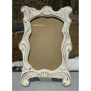 May include: A white ceramic picture frame with ornate gold detailing. The frame has a curved, decorative design with a rectangular opening for a photo. The frame is freestanding and has a vintage aesthetic.