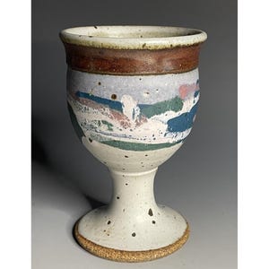 May include: A handmade ceramic goblet with a white stem and base, and a wide, decorated bowl. The bowl features abstract designs in blue, green, and pink against a light blue background, with a brown band at the rim.