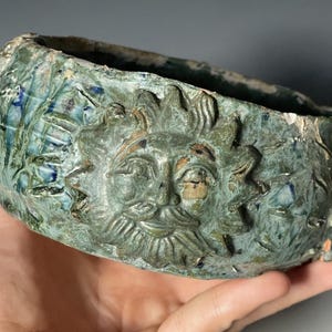 Könnte beinhalten: Eine runde Keramikschale mit einer texturierten, gesprenkelten grün-blauen Glasur. Die Außenseite zeigt ein Reliefdesign eines Sonnengesichts mit Strahlen und stilisierten Blattmustern. Der Rand ist uneben und weist kleine Abplatzungen auf.