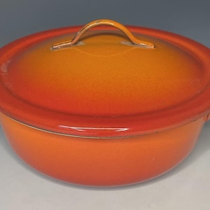 May include: A round, enameled metal casserole dish with a matching lid. The cookware features a vibrant gradient finish that transitions from a deep, saturated orange at the base to a lighter, golden-yellow hue toward the center of the lid.