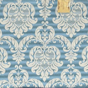May include: A square fabric swatch featuring a repeating white damask pattern over a background of horizontal light and dark blue stripes. A small rectangular tan tag with illegible text is attached to the top center of the fabric piece.