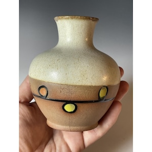 May include: A small, handcrafted ceramic vase with a cream-colored top and a brown base. The base is decorated with a black line and orange and yellow circular accents. The vase is held in a person's hand.