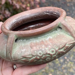 May include: A small, oval-shaped ceramic planter with a brown and green finish. The planter features a floral design and two small holes on the sides. The rim is a darker brown, and the interior is a deep red.