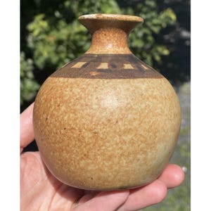 May include: A small, round ceramic vase with a speckled tan body and a dark brown band near the top. The vase has a narrow neck and a flared rim. The design is simple and rustic, suitable for displaying small flowers or as a decorative piece.