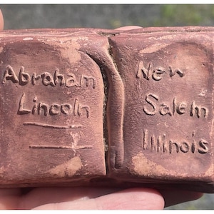 May include: A terracotta-colored book-shaped object with the words "Abraham Lincoln" and two lines on the left side and "New Salem Illinois" on the right side.