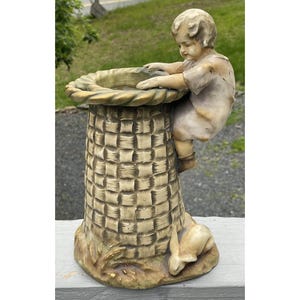 Op de afbeelding: Een vintage tuinbeeld met een jong kind dat op een vogelbad in mandweefstijl klimt. Het beeld is geschilderd in gedempte beige- en bruintinten, met een klein konijn aan de basis. Een decoratief buitenaccent.