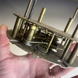 Może przedstawiać: Zbliżenie mechanizmu zegarowego z mosiężnymi kołami zębatymi, sprężynami i metalowymi prętami. Widoczne są skomplikowane wewnętrzne mechanizmy zegara, prezentujące precyzyjną inżynierię urządzenia do pomiaru czasu. Mechanizm zegarowy jest trzymany w dłoni.
