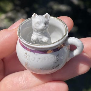 May include: A small, white porcelain teacup with a handle, featuring a white cat figurine inside. The cup has a gold rim and a purple band. The teacup is held in a hand.