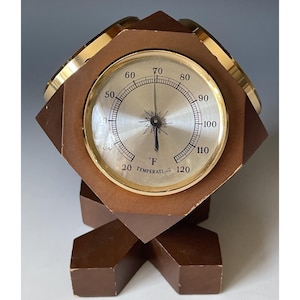 Puede incluir: Un termómetro vintage con un marco de madera marrón y un borde de metal dorado. El termómetro muestra temperaturas en grados Fahrenheit de 20 a 120. La base tiene un diseño geométrico único.