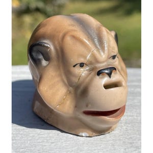 May include: A ceramic piggy bank shaped like a bulldog's head. The piggy bank is painted in shades of brown and beige with black detailing around the eyes and nose. The piggy bank has a slot for coins on the top of the head.