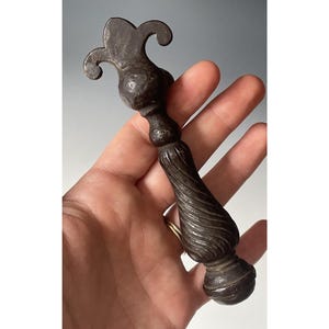 Może przedstawiać: Ciemnobrązowy uchwyt do szuflady w stylu antycznym z ozdobnym wierzchołkiem w kształcie fleur-de-lis i skręconym uchwytem. Metalowy osprzęt jest trzymany w dłoni, prezentując jego skomplikowany design i postarzany wygląd.