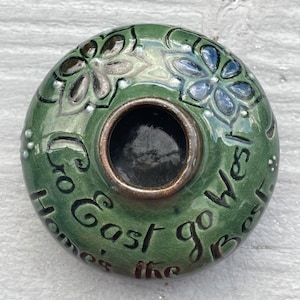 May include: Green ceramic ashtray with a central circular opening and a bronze-colored rim. The ashtray is decorated with floral designs in blue and gray, and the words "Home's the Best, Go East go West" are written around the edge.