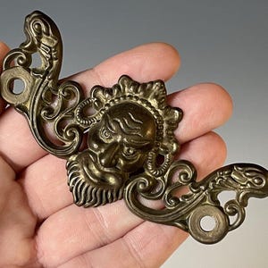 May include: A gold-toned metal decorative piece with a detailed design featuring a stylized face with a beard and intricate scrollwork.