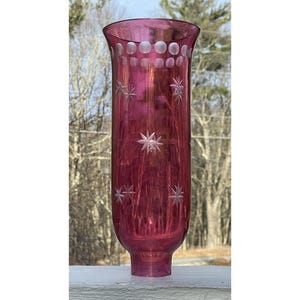 May include: A tall, ruby-coloured glass vase with etched star and dot designs. The vase has a flared top and a slightly bulbous base. The etched stars are evenly spaced along the body of the vase. The vase is approximately 30 cm tall.