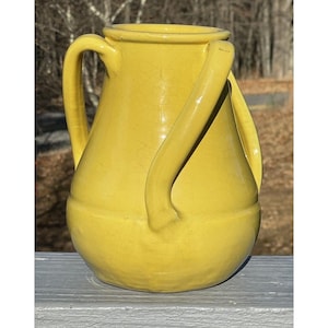 Pode incluir: Um jarro de cerâmica amarelo com duas alças curvas. O jarro tem uma base bulbosa e uma borda ligeiramente alargada. A superfície tem uma textura rachada. O fundo está desfocado, mostrando árvores e uma superfície de madeira.