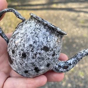 Könnte beinhalten: Eine kleine, schwarz-weiß gesprenkelte Teekanne mit rundem Korpus und einem gebogenen Ausguss. Die Teekanne ist aus Metall und hat einen rustikalen Look.