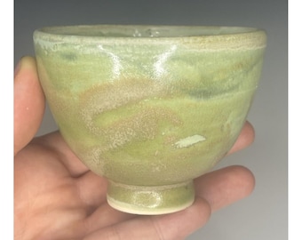 May include: A small, green ceramic bowl with a white glaze and a speckled finish. The bowl has a rounded shape and a wide rim. It is sitting on a hand.