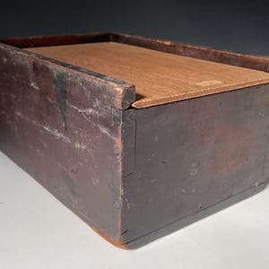 May include: A rectangular wooden box with a dark brown, weathered exterior finish and a lighter, natural-toned wood sliding lid. The box features visible joinery at the corners and a smooth, flat top surface that fits into grooves along the sides.
