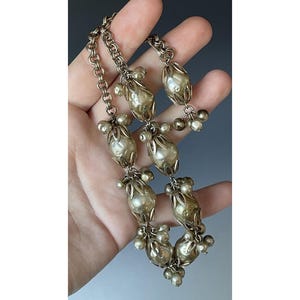 Puede incluir: Un collar vintage plateado con eslabones ovalados ornamentados y detalles de perlas. El collar está compuesto por múltiples hilos y pequeños grupos de perlas colgantes, creando un diseño delicado y elegante.