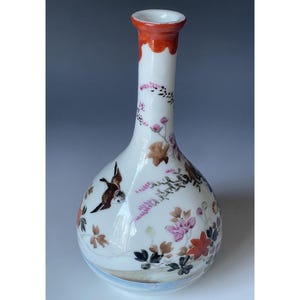 May include: A white ceramic vase with a bulbous body and a narrow neck. The rim is orange, and the vase is decorated with floral and bird motifs in shades of brown, pink, and black. The vase is approximately 10 inches tall.