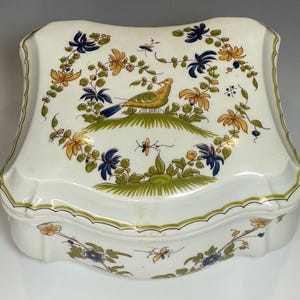 Könnte beinhalten: Eine weiße Porzellan-Schmuckdose mit einem gewellten Rand. Der Deckel ist mit einem floralen Muster in Blau-, Gelb- und Grüntönen verziert, auf dem ein Vogel auf einem Ast sitzt.