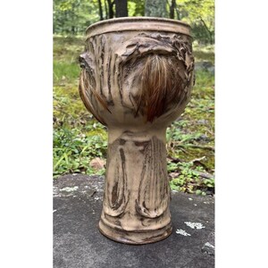 May include: A ceramic vase with a textured, brown and beige finish. The vase has a wide, round opening and a tall, slender base. The vase is decorated with a raised design of leaves and branches.